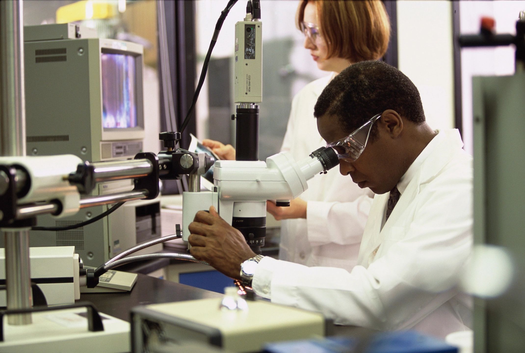 A black male scientist looking in a microscope in a lab with a white female scientist in the background